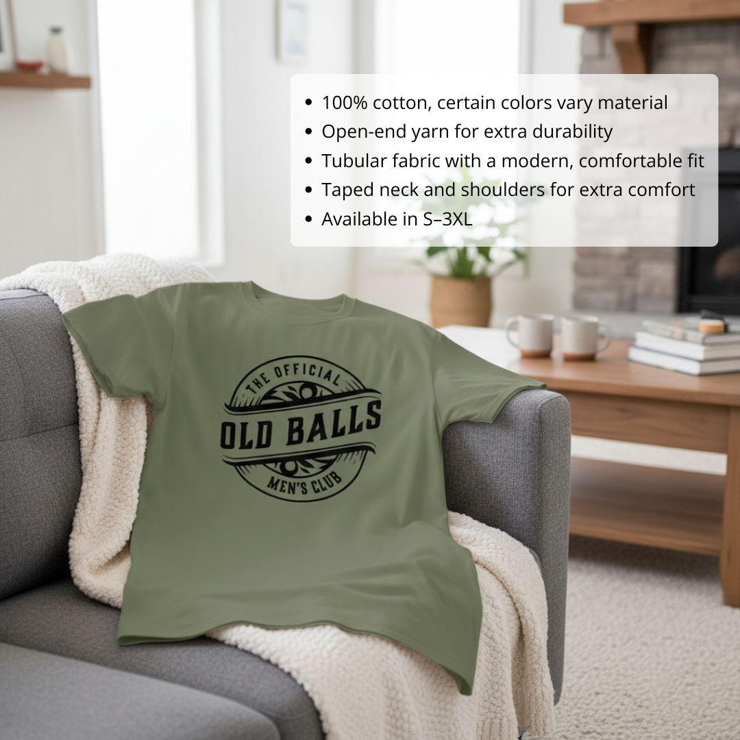 Sage green cotton t-shirt with "The Official Old Balls Men's Club" printed in black circular badge design, displayed on gray sofa with blanket throw