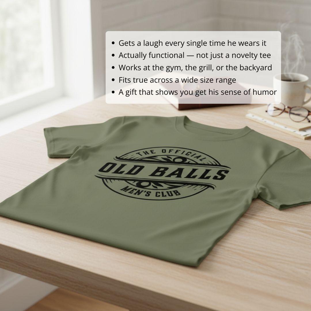 Sage green t-shirt with black printed graphic reading "The Official Old Balls Men's Club" laid flat on wooden table with lifestyle styling