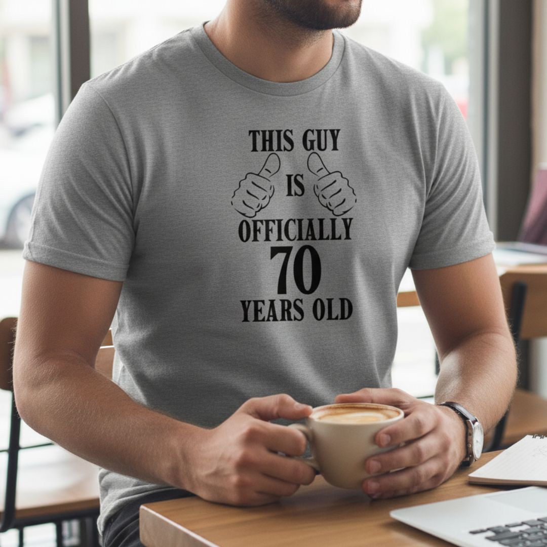 Gray t-shirt printed with "This Guy is Officially 70 Years Old" text and dual thumbs-up graphic, shown on model holding coffee cup
