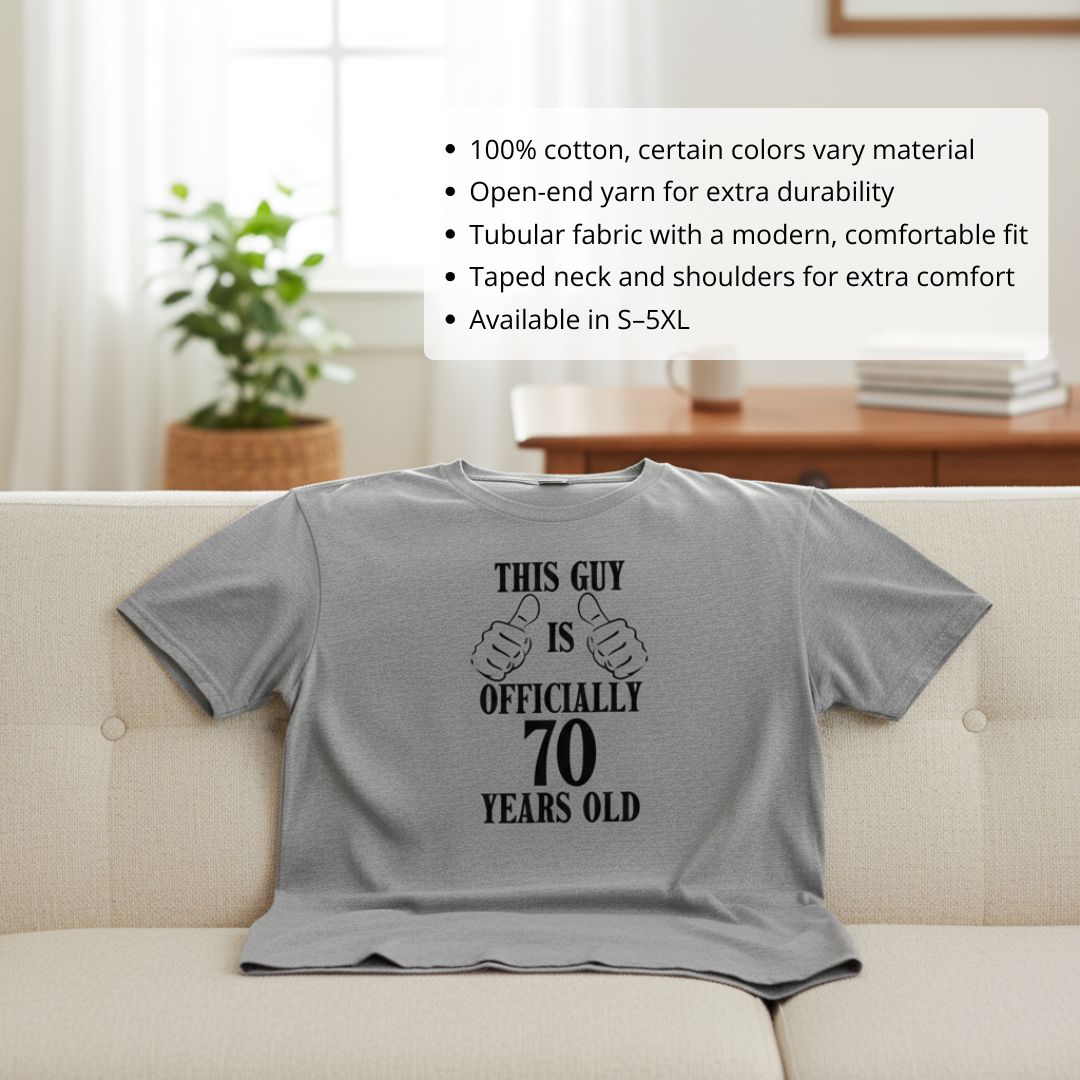 Gray t-shirt with black printed text reading "This Guy is Officially 70 Years Old" with thumbs-up graphics, displayed on cream-colored couch