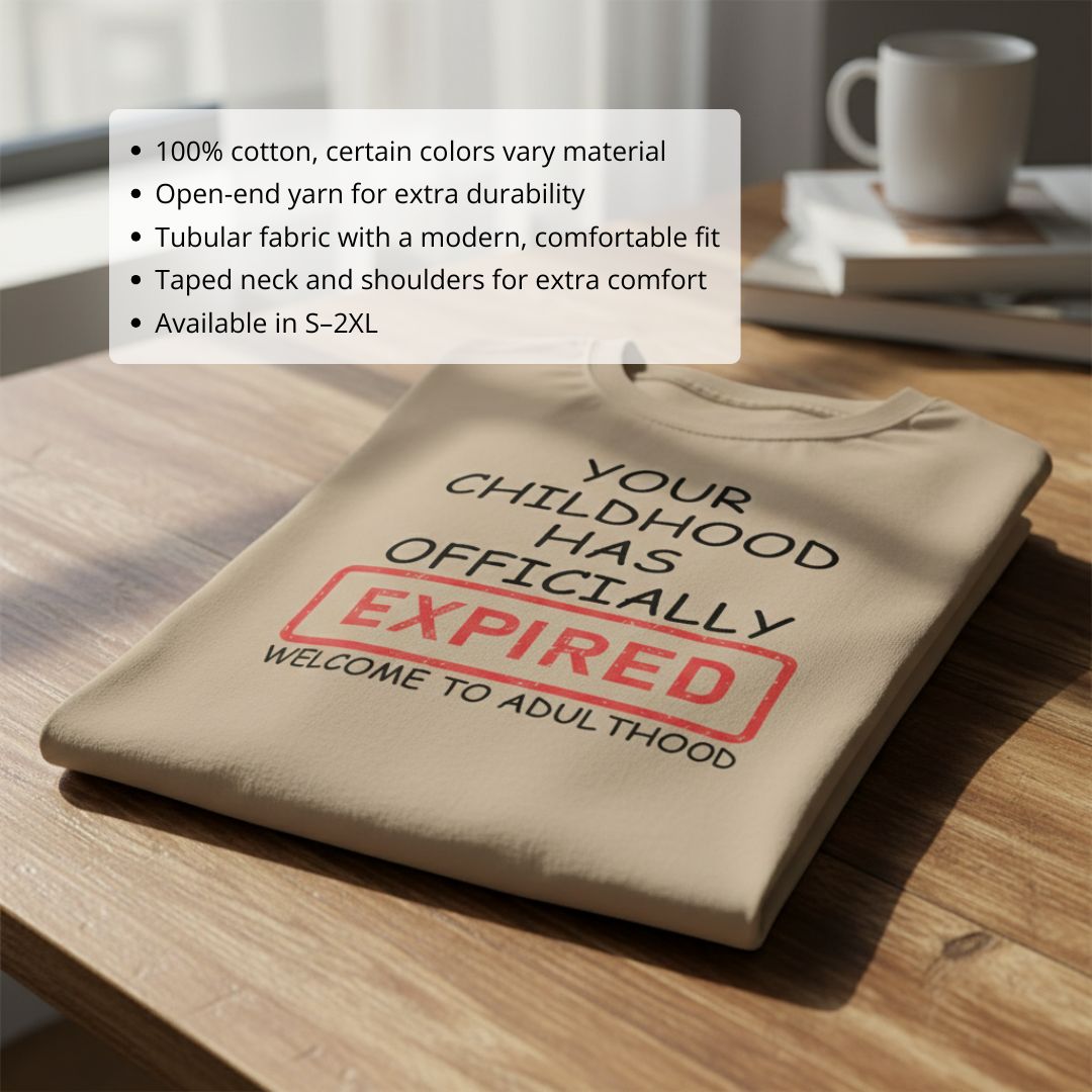 Tan t-shirt with screen-printed text reading "Your Childhood Has Officially Expired" with red stamp graphic and "Welcome to Adulthood" below, folded on wooden surface with mug and books in background.