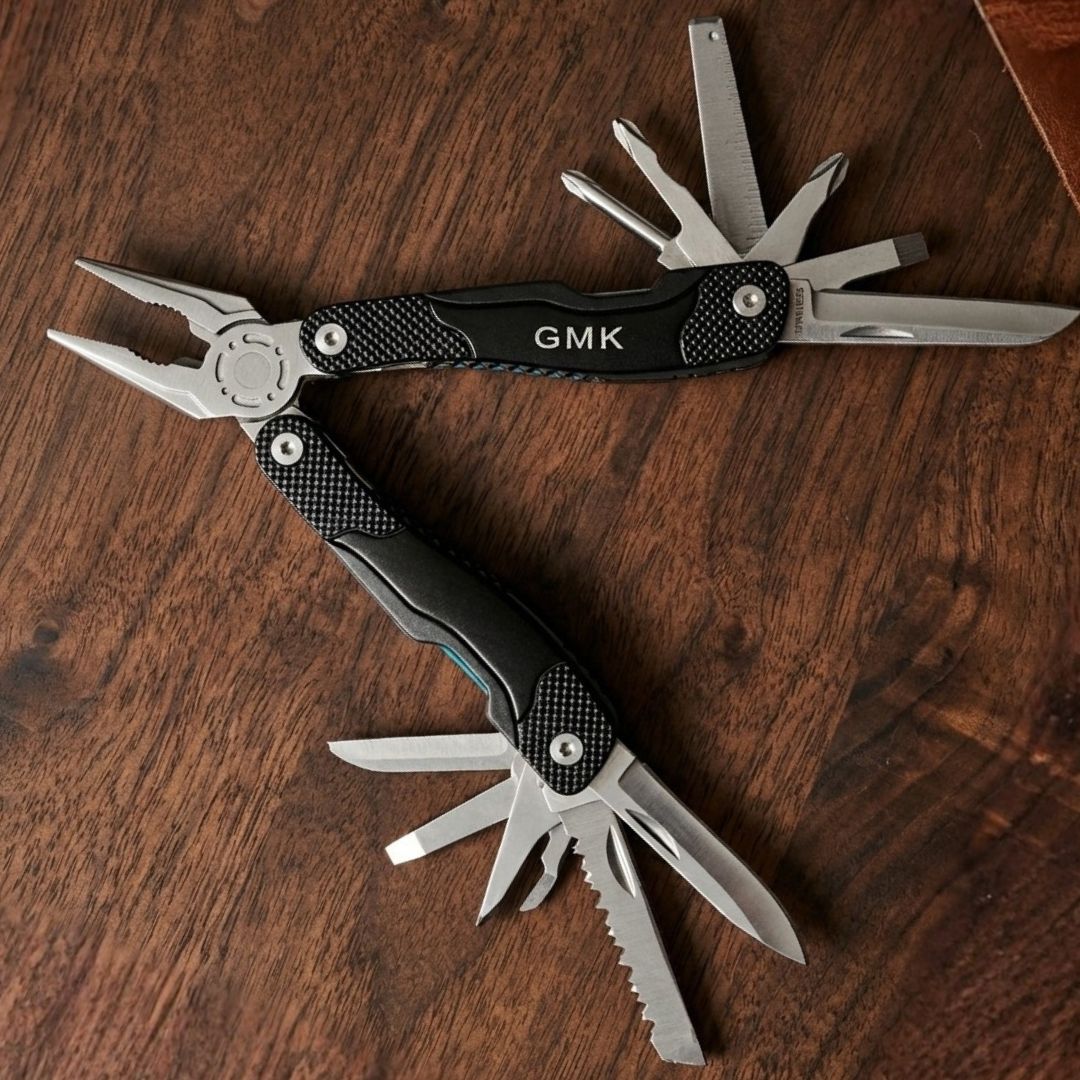 Black multi-tool with carbon fiber handles engraved with sample initials "GMK", displayed open showing stainless steel blades and tools on wooden surface.