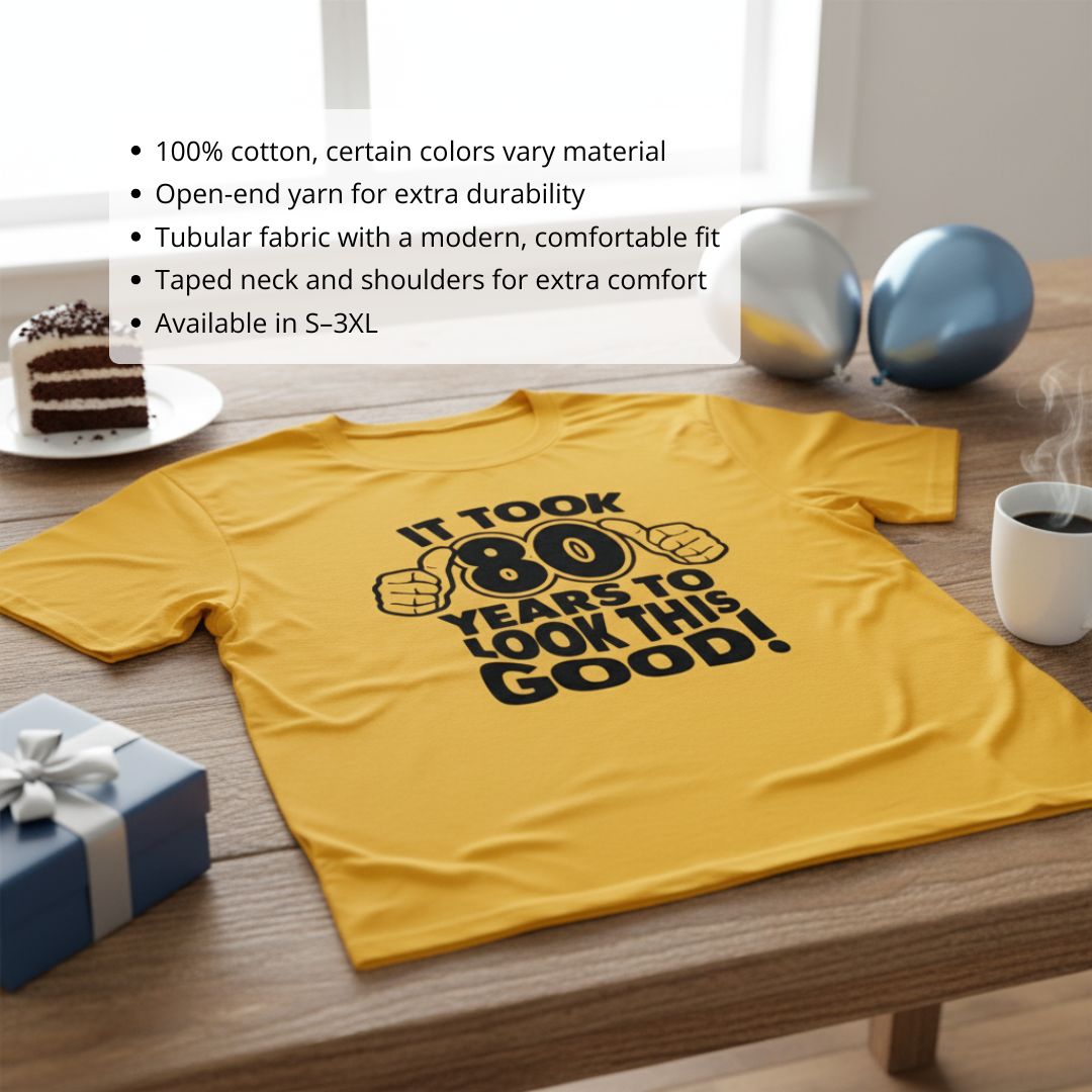 Gold t-shirt printed with "It took 80 years to look this good" text and thumbs-up graphic design, laid flat on wooden surface.