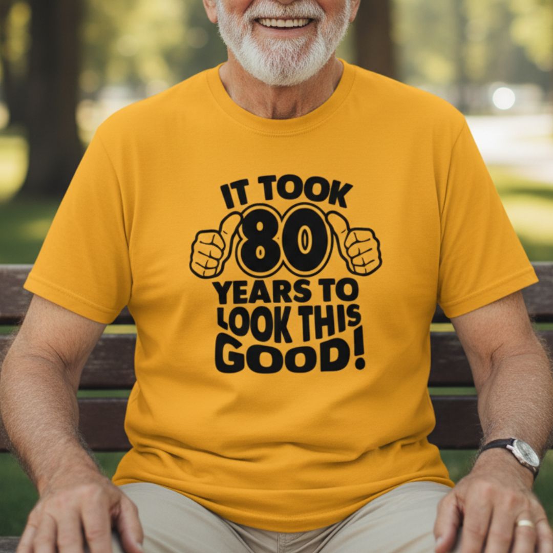Gold t-shirt with black text and thumbs-up graphics reading "It took 80 years to look this good" worn by man outdoors