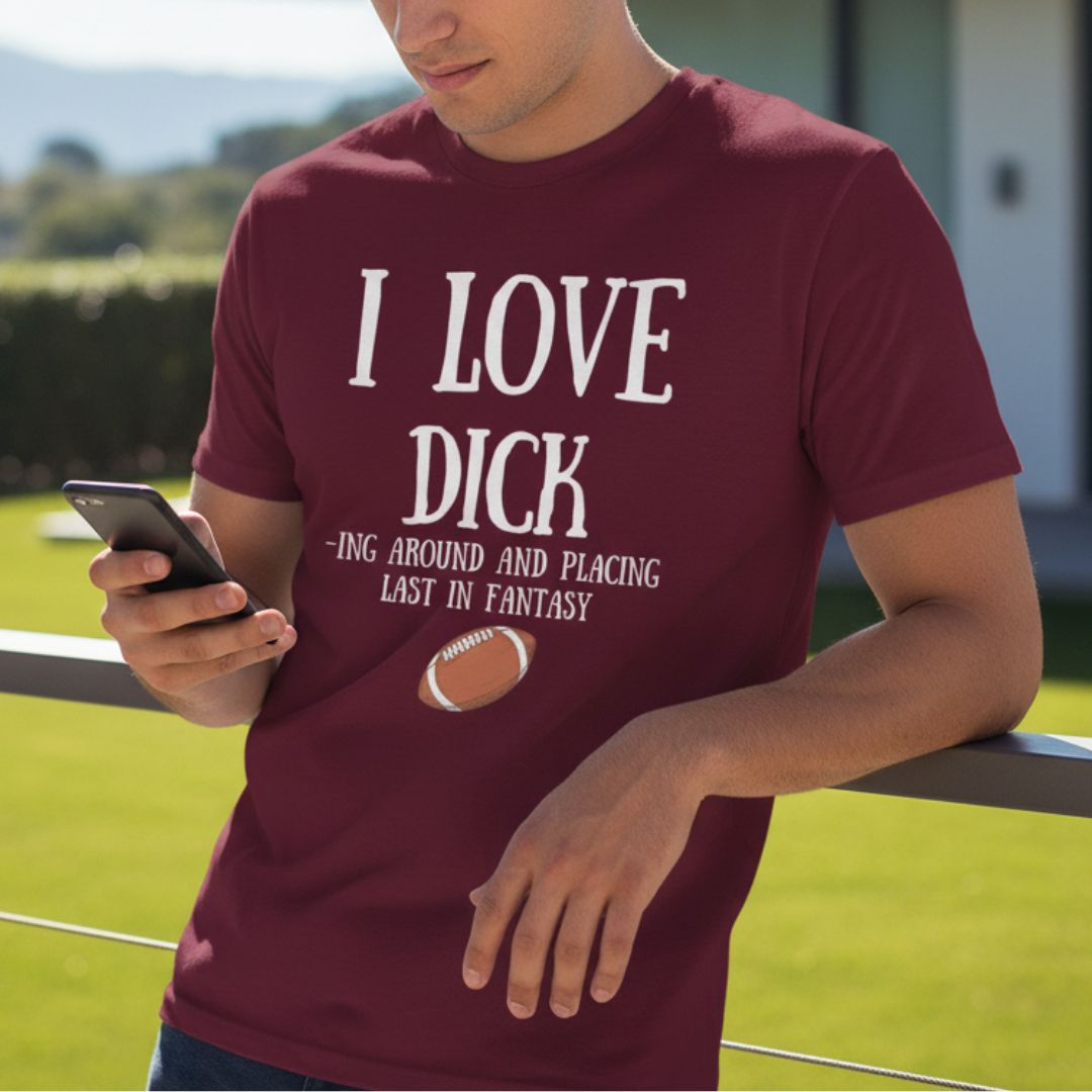 Maroon t-shirt printed with "I LOVE DICK -ING AROUND AND PLACING LAST IN FANTASY" text and football graphic, worn by man holding phone outdoors