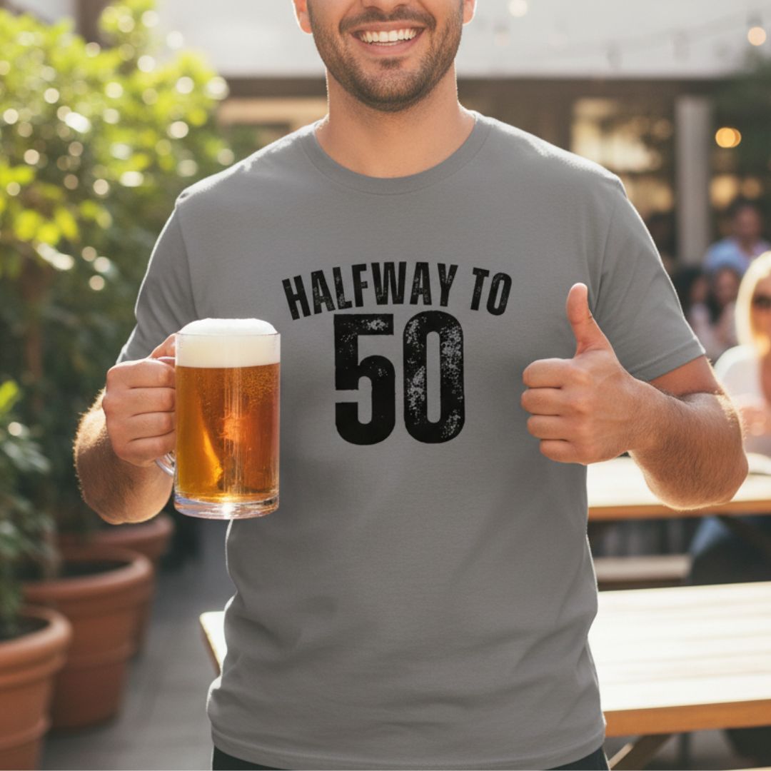 Gray unisex t-shirt printed with black text reading "Halfway to 50" worn by man holding beer mug outdoors