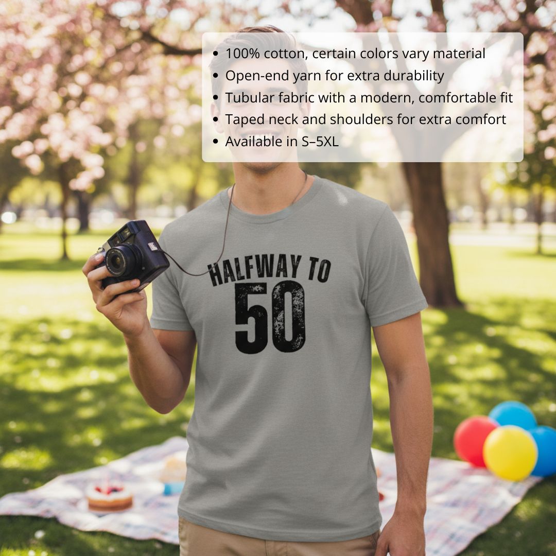 Man wearing gray cotton t-shirt printed with "HALFWAY TO 50" text, holding vintage camera outdoors in park setting with balloons visible
