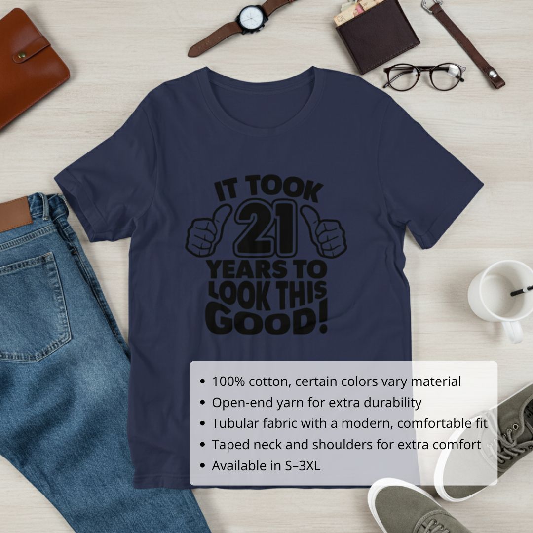 Navy crewneck t-shirt with black graphic reading "It took 21 years to look this good" with thumbs-up design, displayed flat with accessories including watch, wallet, and glasses