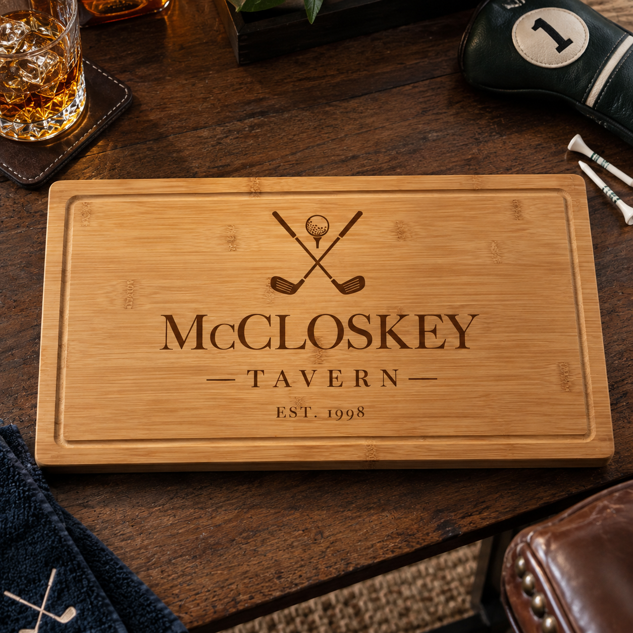 Angled view of an engraved bamboo golf cutting board reading McCloskey Tavern, displayed beside a whiskey glass, golf club headcover, and tees on a wood table.