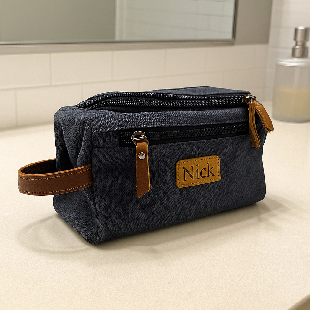 Navy canvas toiletry bag with brown leather handles and trim, personalized with sample engraving "Nick" on tan leather patch, displayed on bathroom vanity