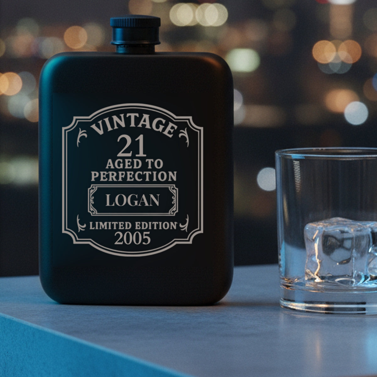 Personalized 21st birthday flask engraved with the name Logan and the year 2005, on an apartment kitchen island at night with city lights behind.