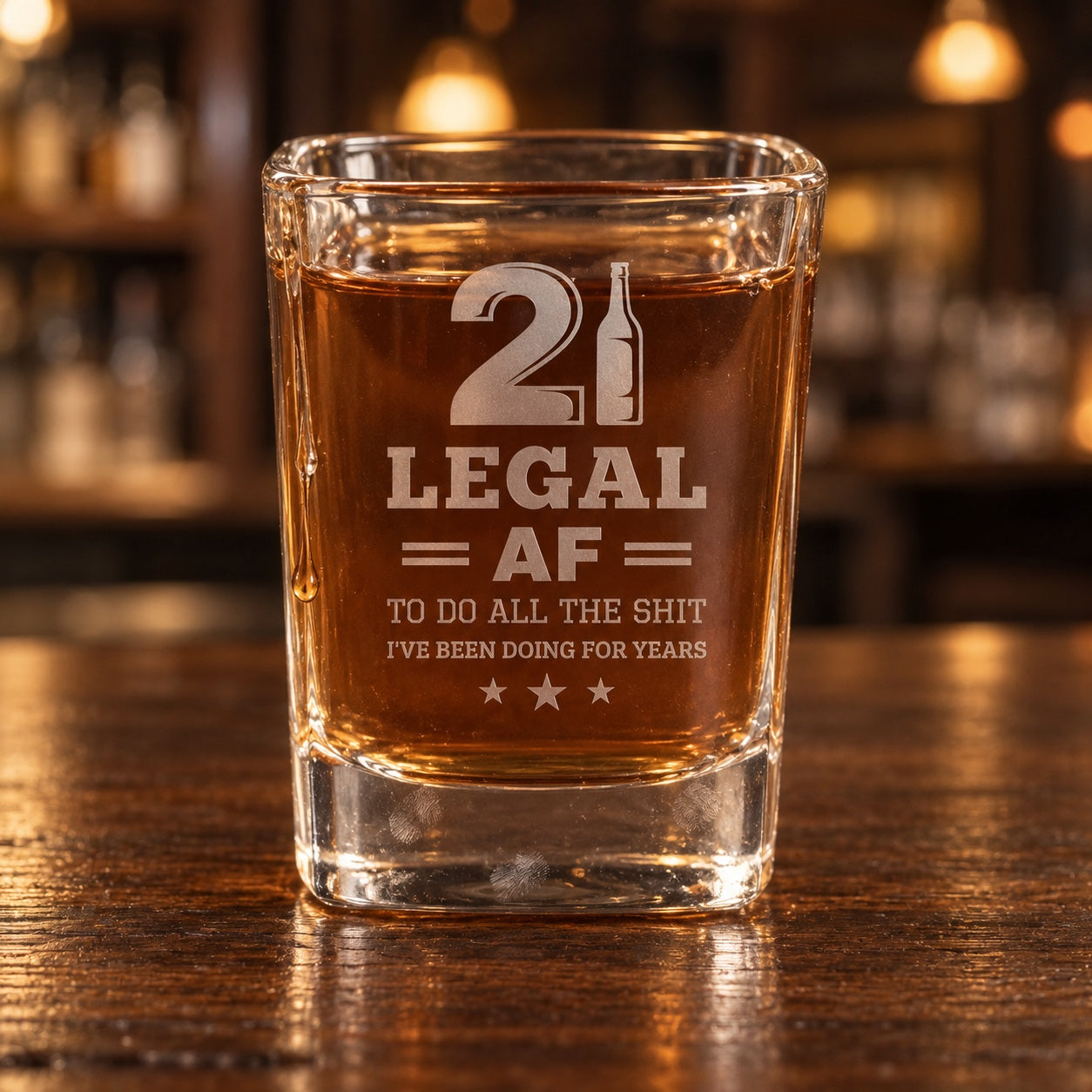 21st birthday shot glass filled with whiskey on a walnut bar, engraved with a big 21, LEGAL AF and a funny line for his first legal shot