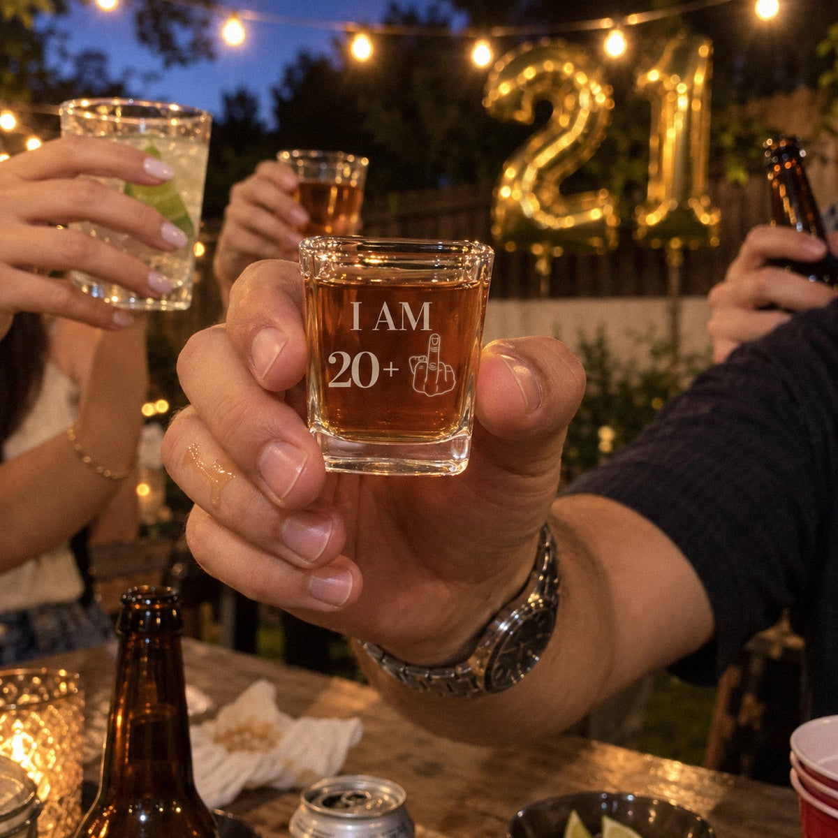 A friend raises an I AM 20 plus shot glass at a 21st birthday party in the backyard, gold 21 balloons and string lights behind