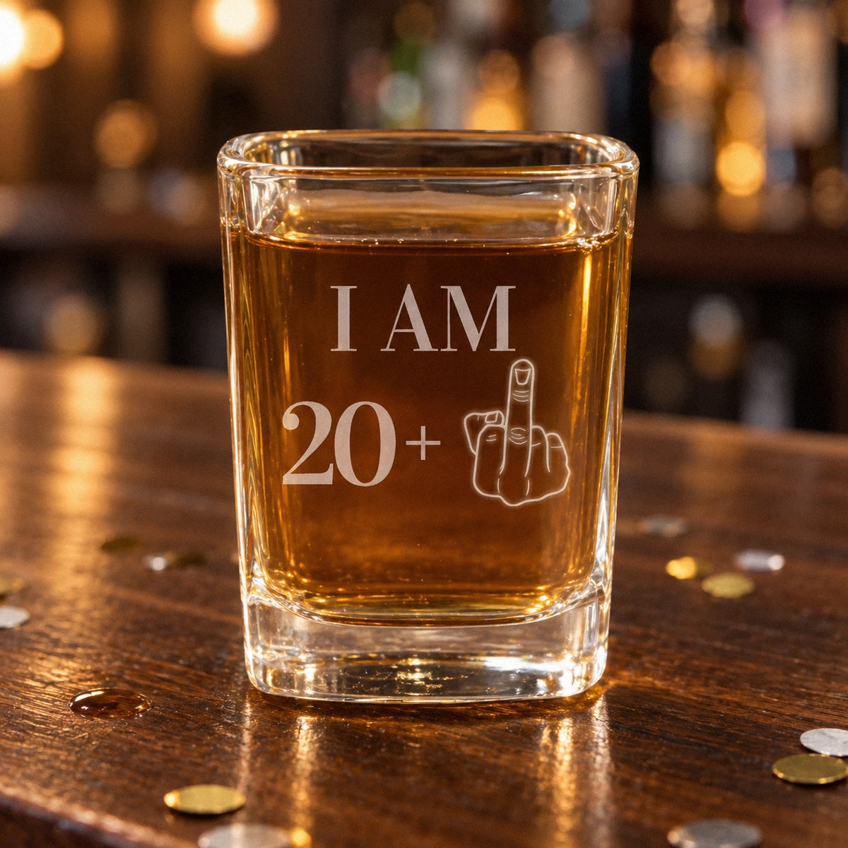 Funny 21st birthday shot glass engraved I AM 20 plus a middle finger, filled with whiskey on a dark walnut bar with confetti