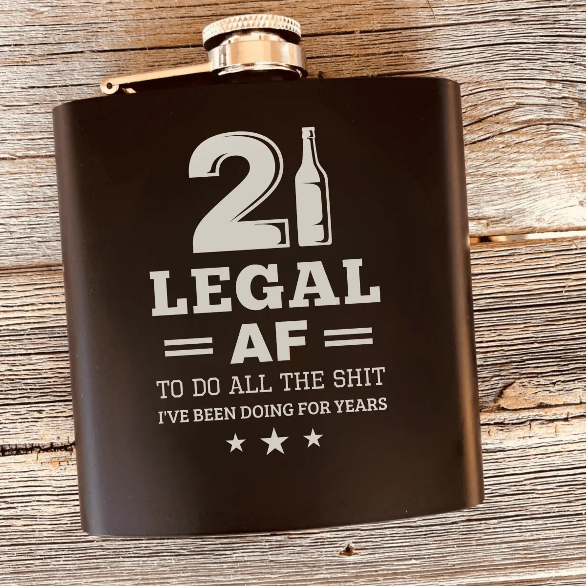 Black stainless steel flask with silver engraved text reading "21 Legal AF to do all the SH*T I've been doing for years" and beer bottle icon with stars, displayed on wooden surface.