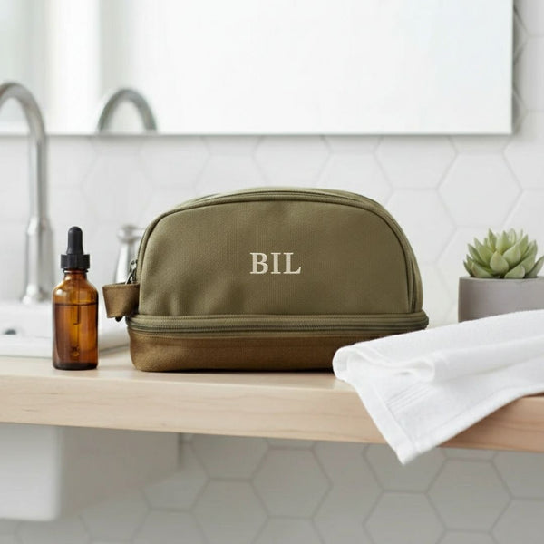 Personalized olive green canvas toiletry bag embroidered with the initials BIL in cream thread, standing on a bathroom vanity shelf