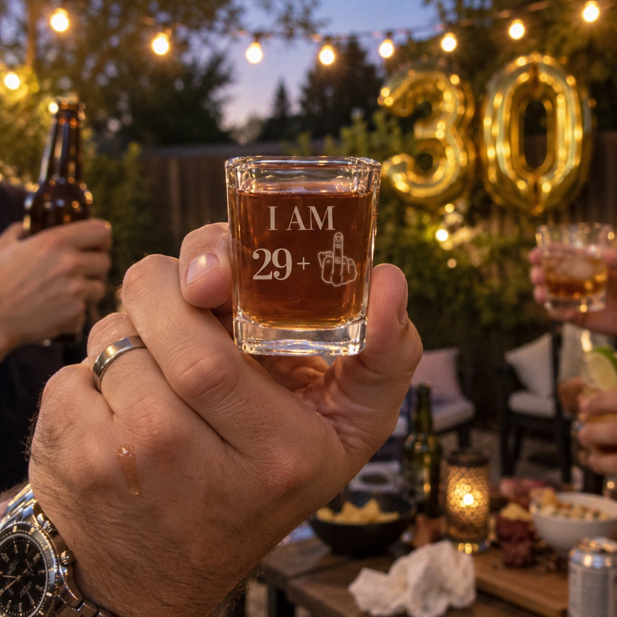 A man raises an engraved I AM 29 plus shot glass at his 30th birthday party in the backyard, gold 30 balloons behind him