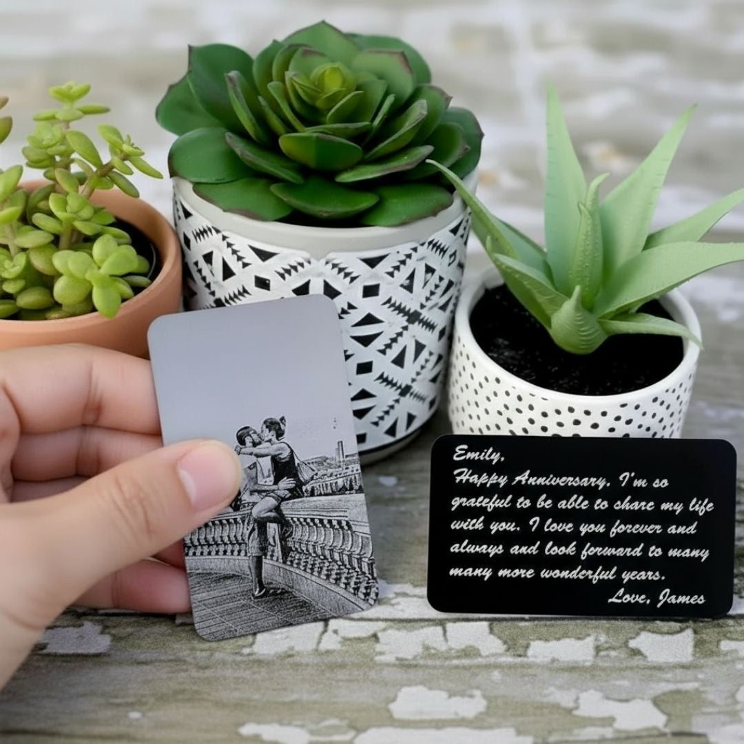 Hand holding personalized wallet insert card with laser-engraved couple photo, black anodized aluminum with sample anniversary message from James on reverse side, displayed with decorative succulents