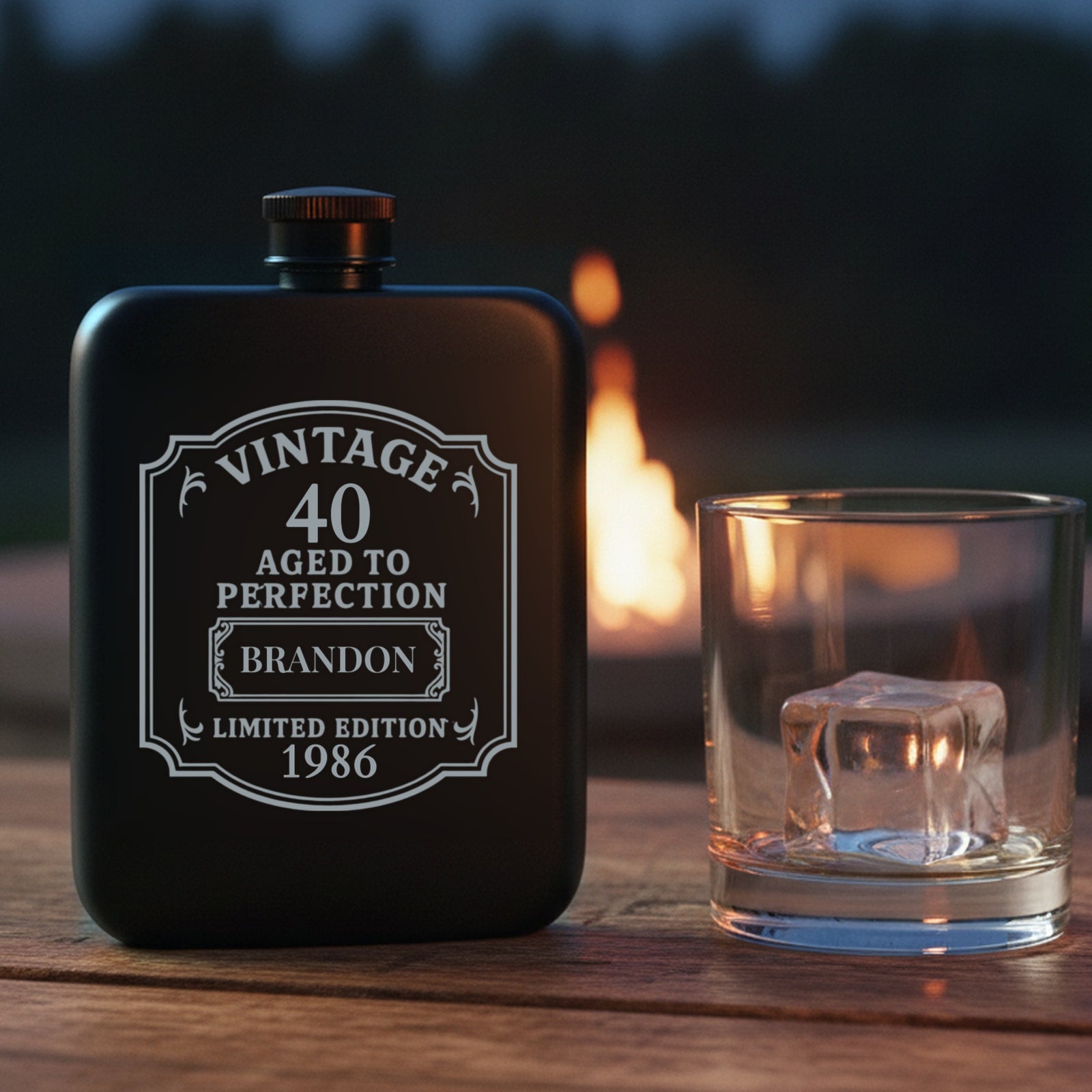 Personalized 40th birthday flask engraved with the name Brandon and the year 1986, on a backyard patio table at dusk beside a lit stone fire pit.