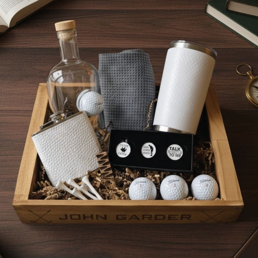 Wooden gift crate personalized with "JOHN GARDER" containing whiskey decanter with golf ball, white tumbler, golf balls, towels, ball markers, and tees on dark wood surface