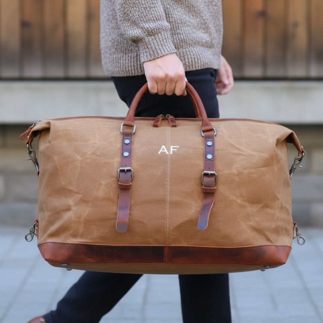 Tan waxed canvas duffle bag with brown leather handles and trim, personalized with sample monogram "AF" embroidered on front, held by person wearing gray sweater