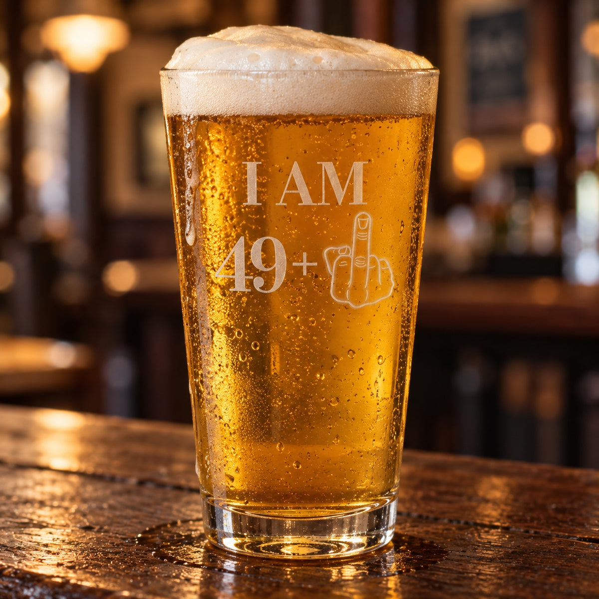 50th birthday beer glass etched I AM 49+ with a middle-finger hand, a 16 oz pint of golden lager on a dark wood bar in a warm pub