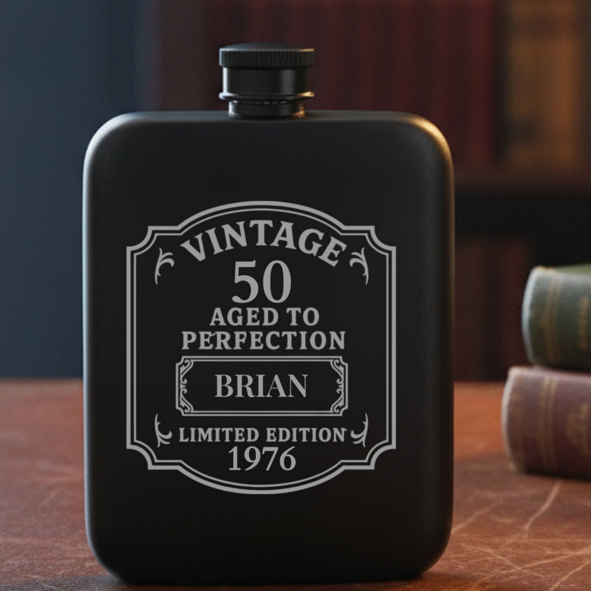 Personalized 50th birthday flask engraved with the name Brian and the year 1976, on a leather study desk beside old clothbound books and reading glasses.
