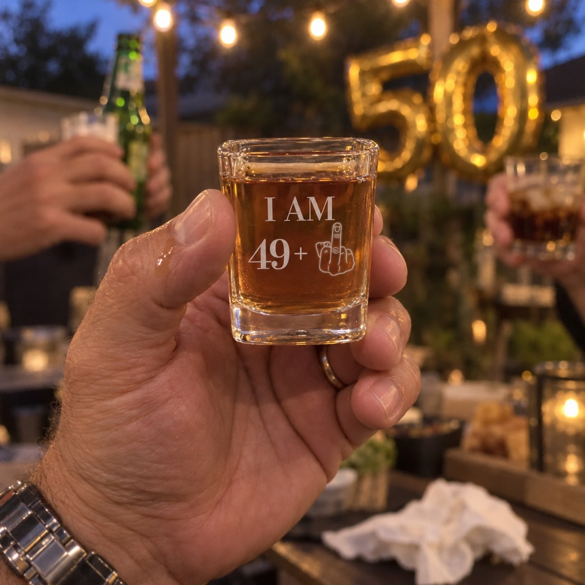 A man raises an engraved I AM 49 plus shot glass at his 50th birthday party in the backyard, gold 50 balloons behind him