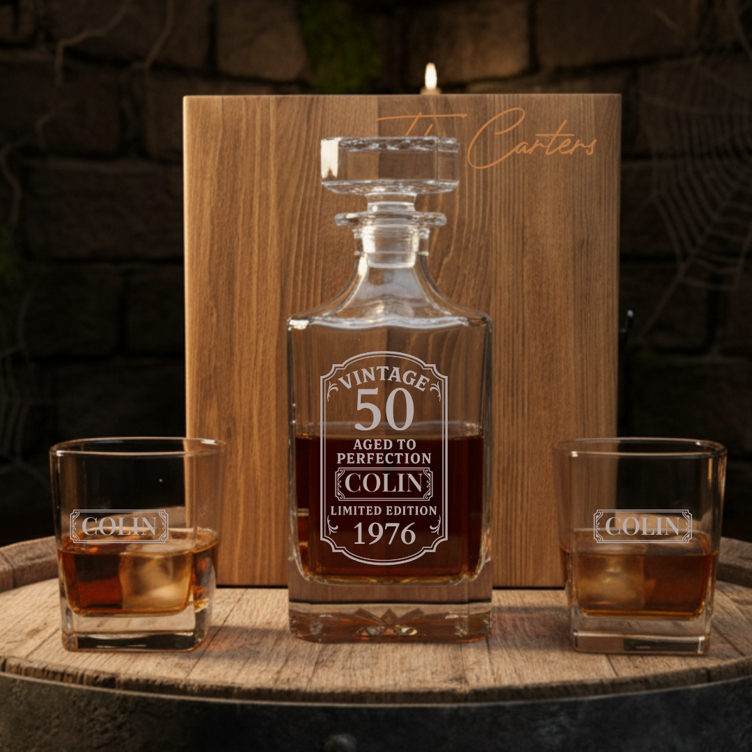 Personalized birthday whiskey decanter set engraved for a 50th birthday, shown with two matching rocks glasses on a barrel