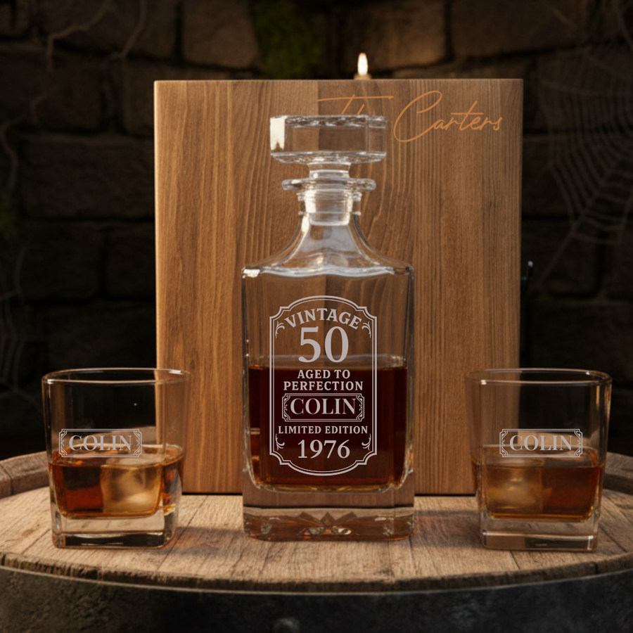 Personalized birthday whiskey decanter set engraved for a 50th birthday, shown with two matching rocks glasses on a barrel