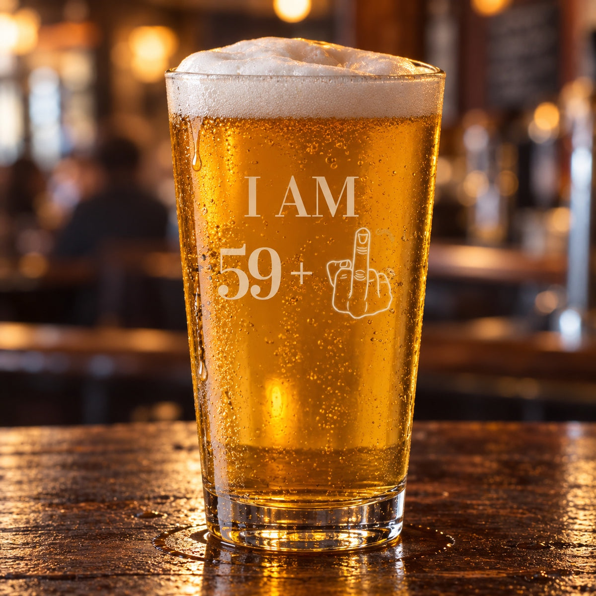 60th birthday beer glass etched I AM 59+ with a middle-finger hand, a 16 oz pint of golden lager on a dark wood bar in a warm pub