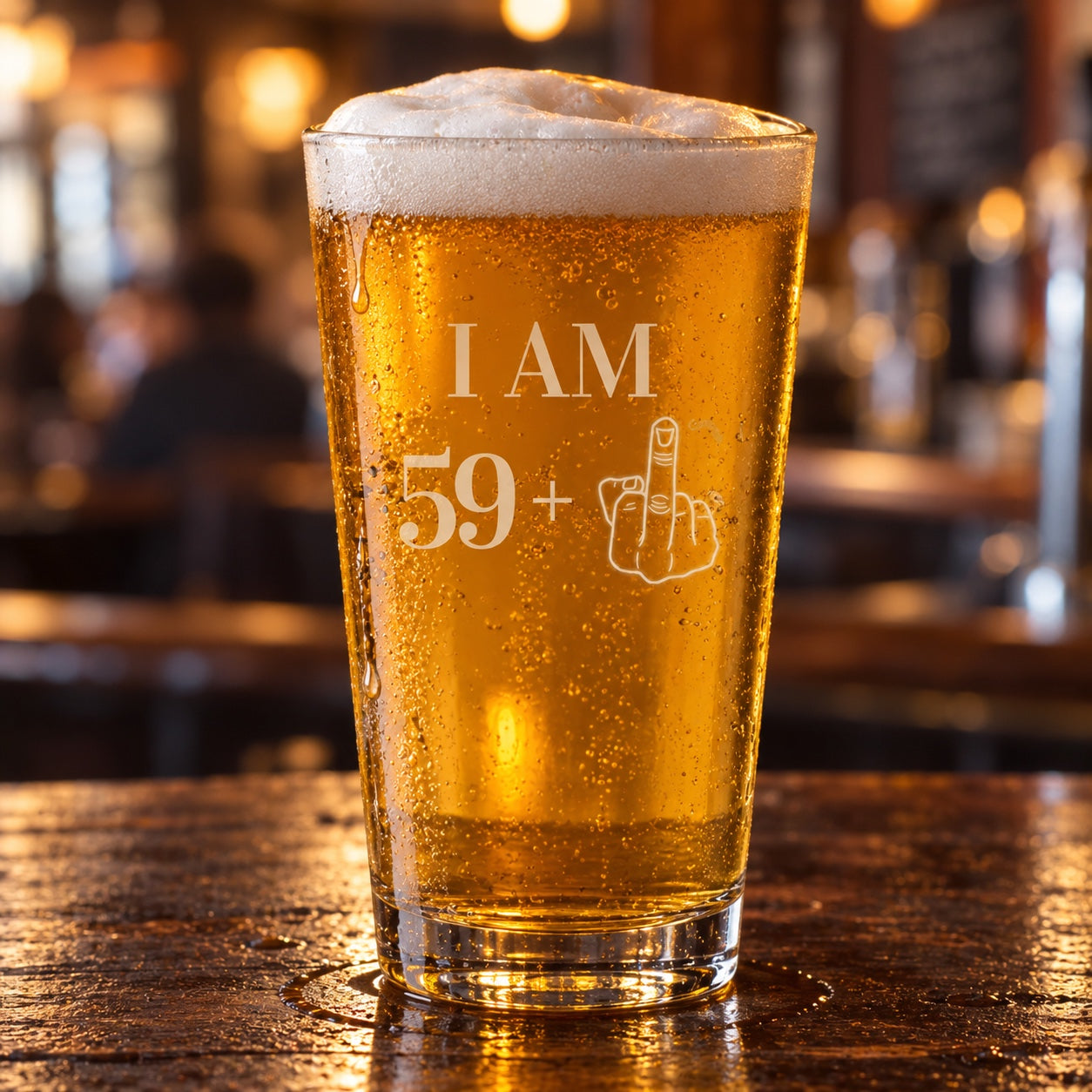 60th birthday beer glass etched I AM 59+ with a middle-finger hand, a 16 oz pint of golden lager on a dark wood bar in a warm pub