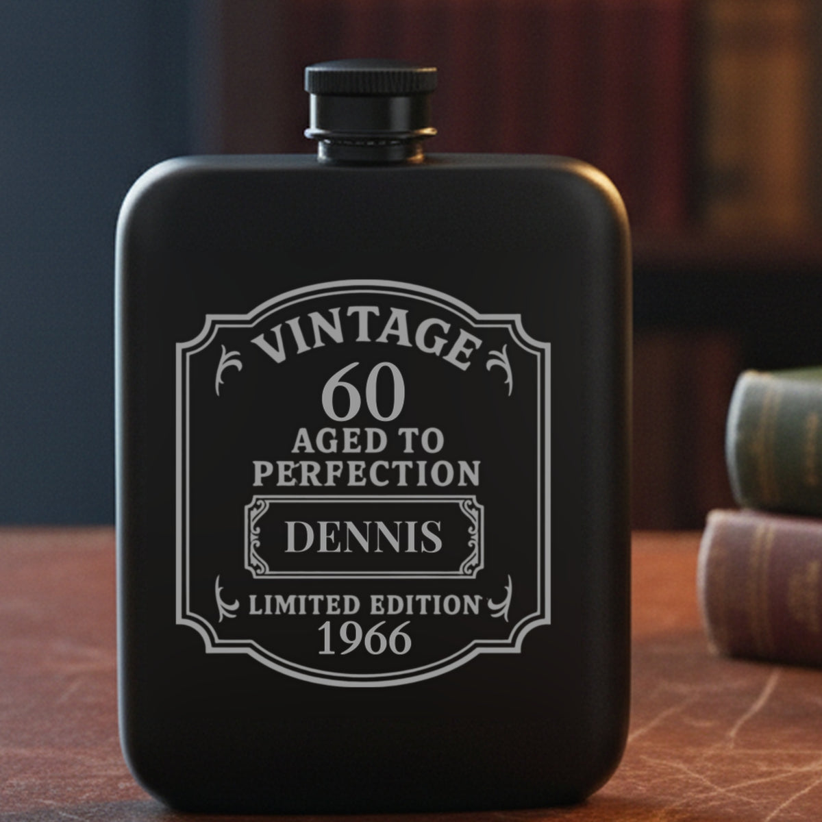 Personalized 60th birthday flask engraved with the name Dennis and the year 1966, on a leather study desk beside old clothbound books and reading glasses.