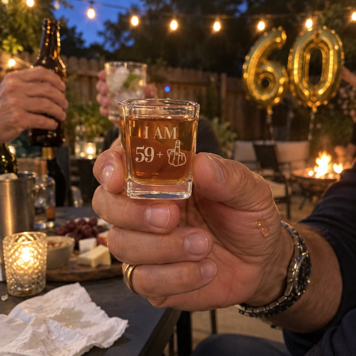 A man raises his I AM 59 plus engraved shot glass in a toast at his 60th birthday party in the backyard, gold 60 balloons and string lights behind.