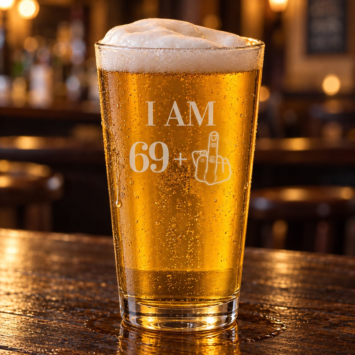 70th birthday beer glass etched I AM 69+ with a middle-finger hand, a 16 oz pint of golden lager on a dark wood bar in a warm pub
