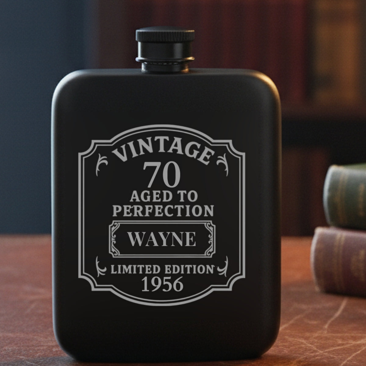 Personalized 70th birthday flask engraved with the name Wayne and the year 1956, on a leather study desk beside old clothbound books and reading glasses.