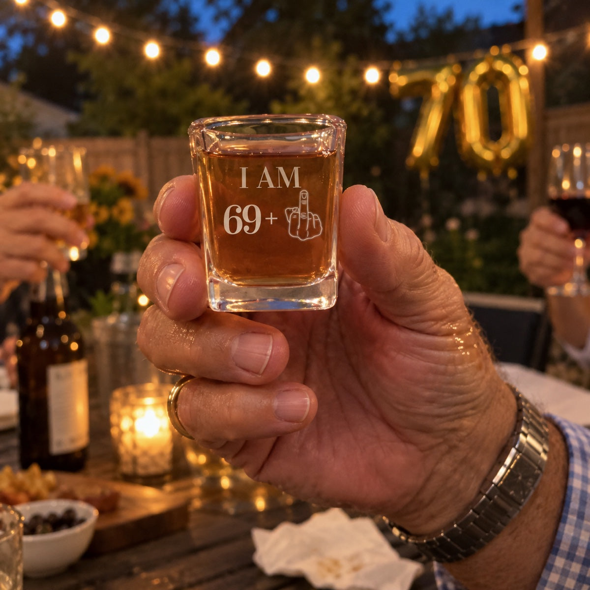 A man raises an engraved I AM 69 plus shot glass at his 70th birthday party in the backyard, gold 70 balloons behind him