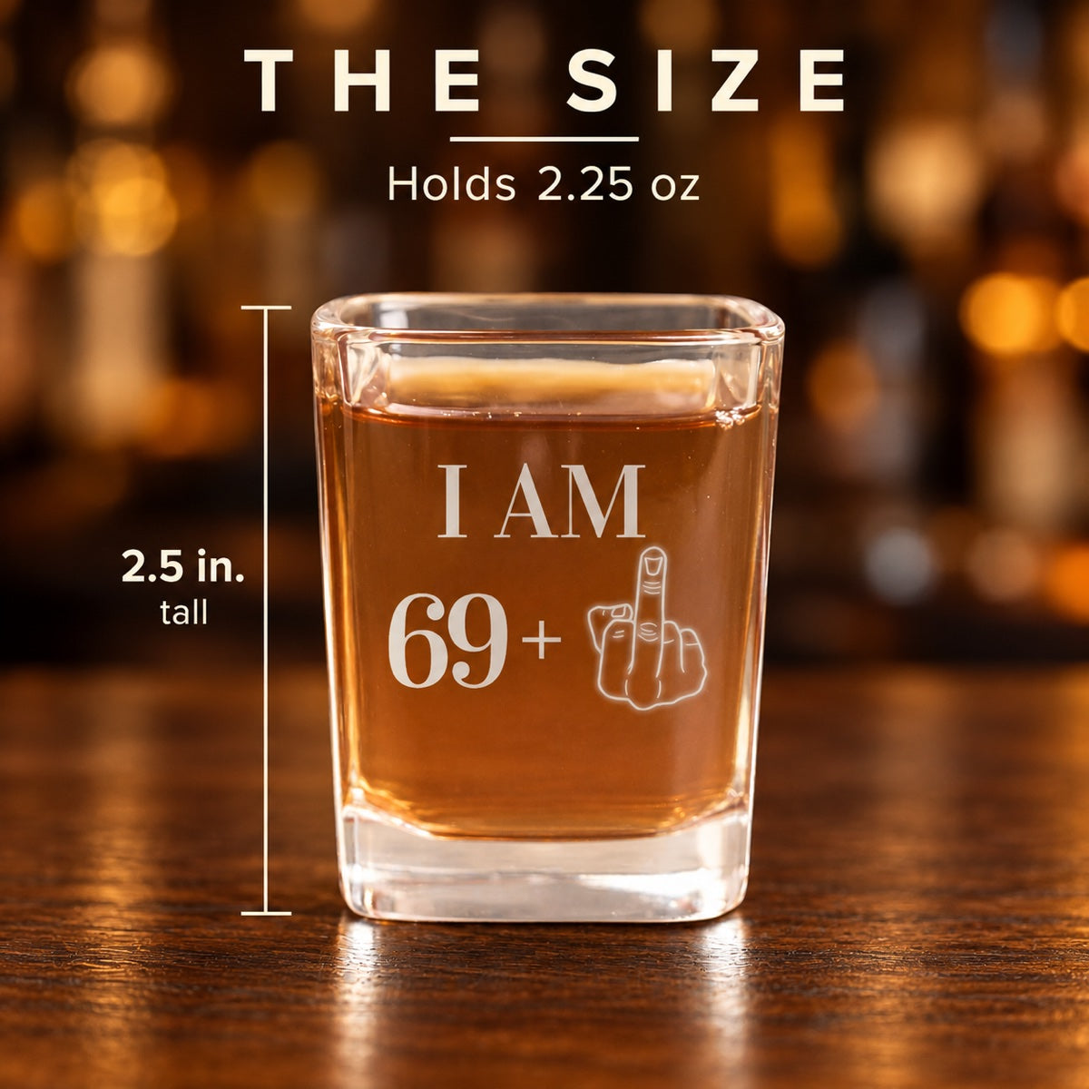 Size of the 70th shot glass engraved I AM 69 plus a middle finger: 2.5 inches tall and holds 2.25 ounces, shown on a walnut bar