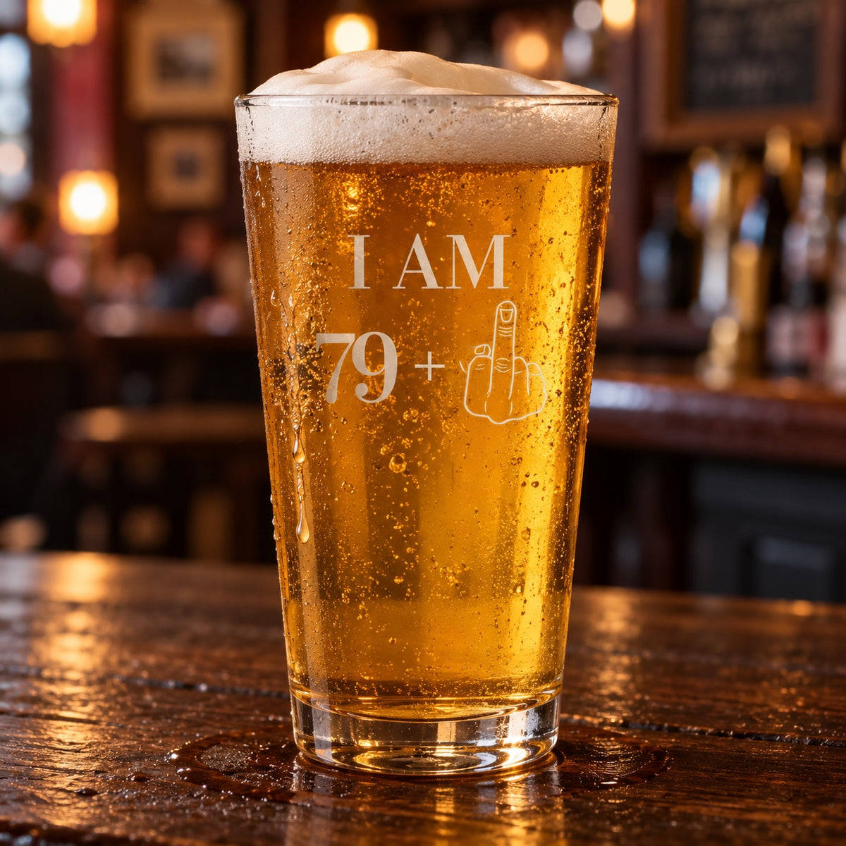 80th birthday glass etched I AM 79+ with a middle-finger hand, a 16 oz pint of golden lager on a dark wood bar in a warm pub