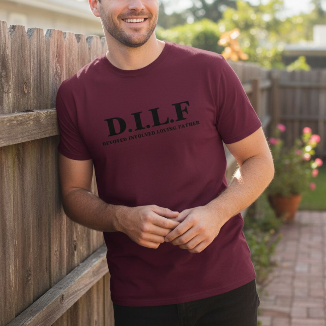 Man wearing maroon t-shirt printed with "D.I.L.F. DEVOTED INVOLVED LOVING FATHER" text, standing against wooden fence in backyard setting