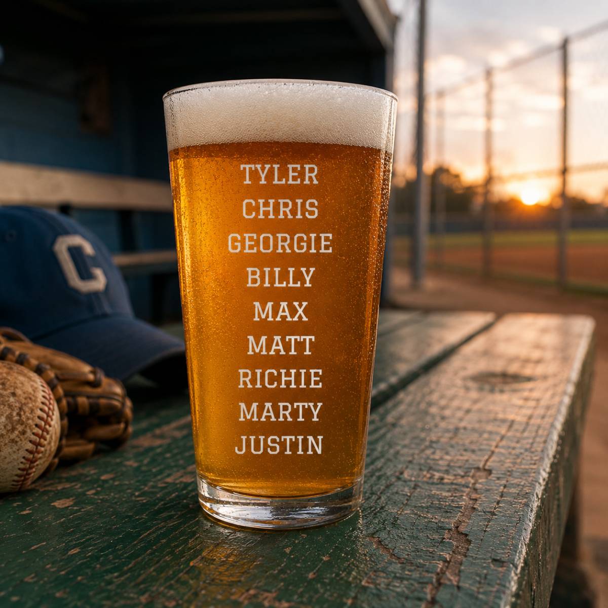 Engraved pint glass with sample names Tyler, Chris, Georgie, Billy, Max, Matt, Richie, Marty, and Justin etched vertically, filled with beer and positioned on a dugout bench at a baseball field at sunset.