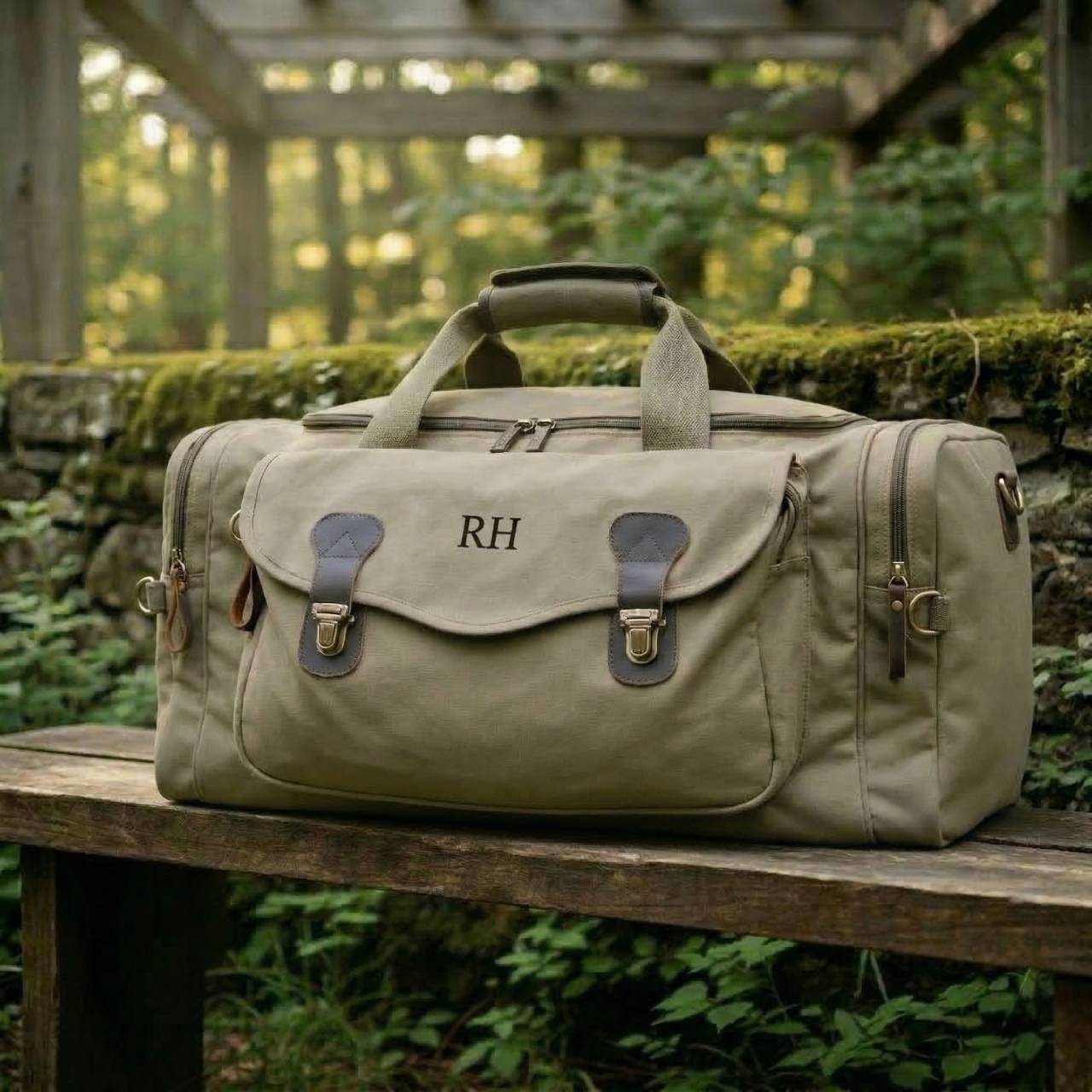 Tan canvas weekender bag with leather accents and handles, personalized with sample monogram "RH" on flap, displayed on wooden bench in garden setting