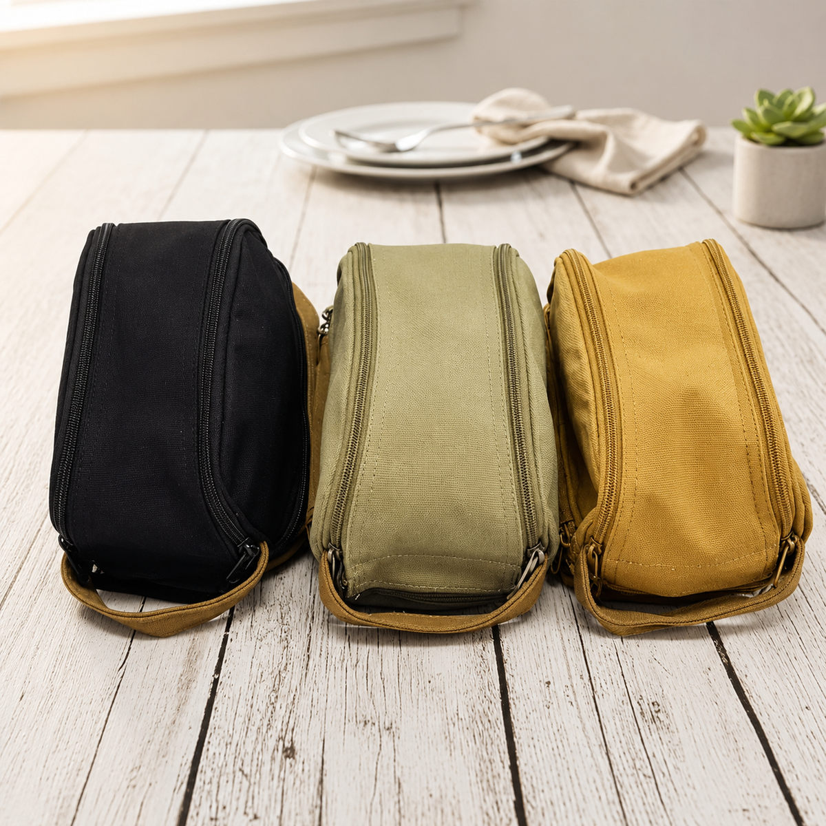 Three canvas toiletry bags in black, sage green, and gold with leather trim and top handles arranged on a white wood surface