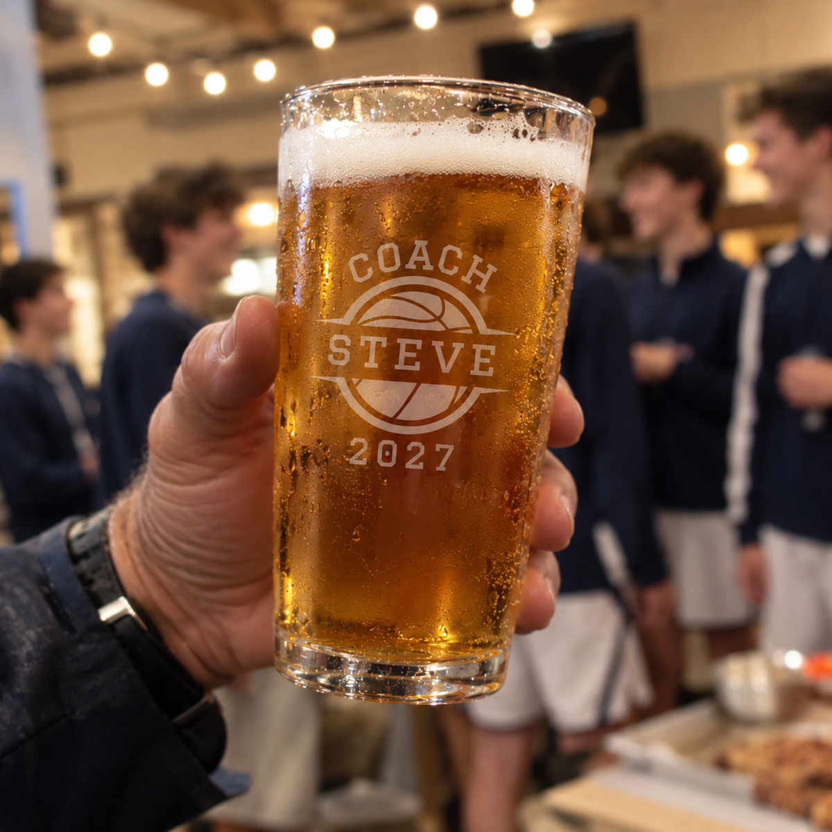 Pint glass filled with beer, engraved with sample text "Coach Steve" and basketball icon with year "2027", held in hand at an event