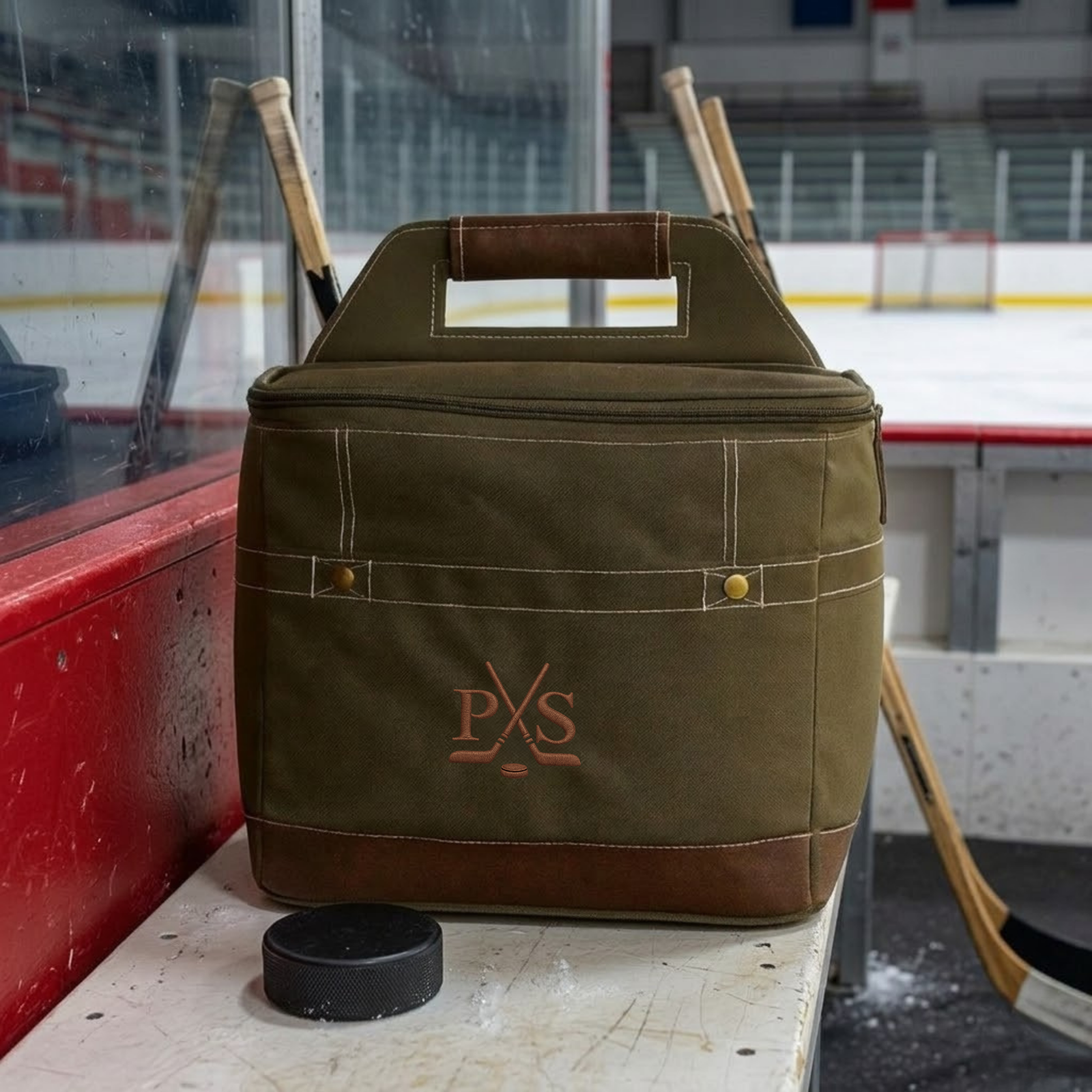Olive canvas and brown leather hockey cooler with dual handles and monogrammed initials "PKS" in copper, positioned on ice rink bench with hockey stick and puck nearby