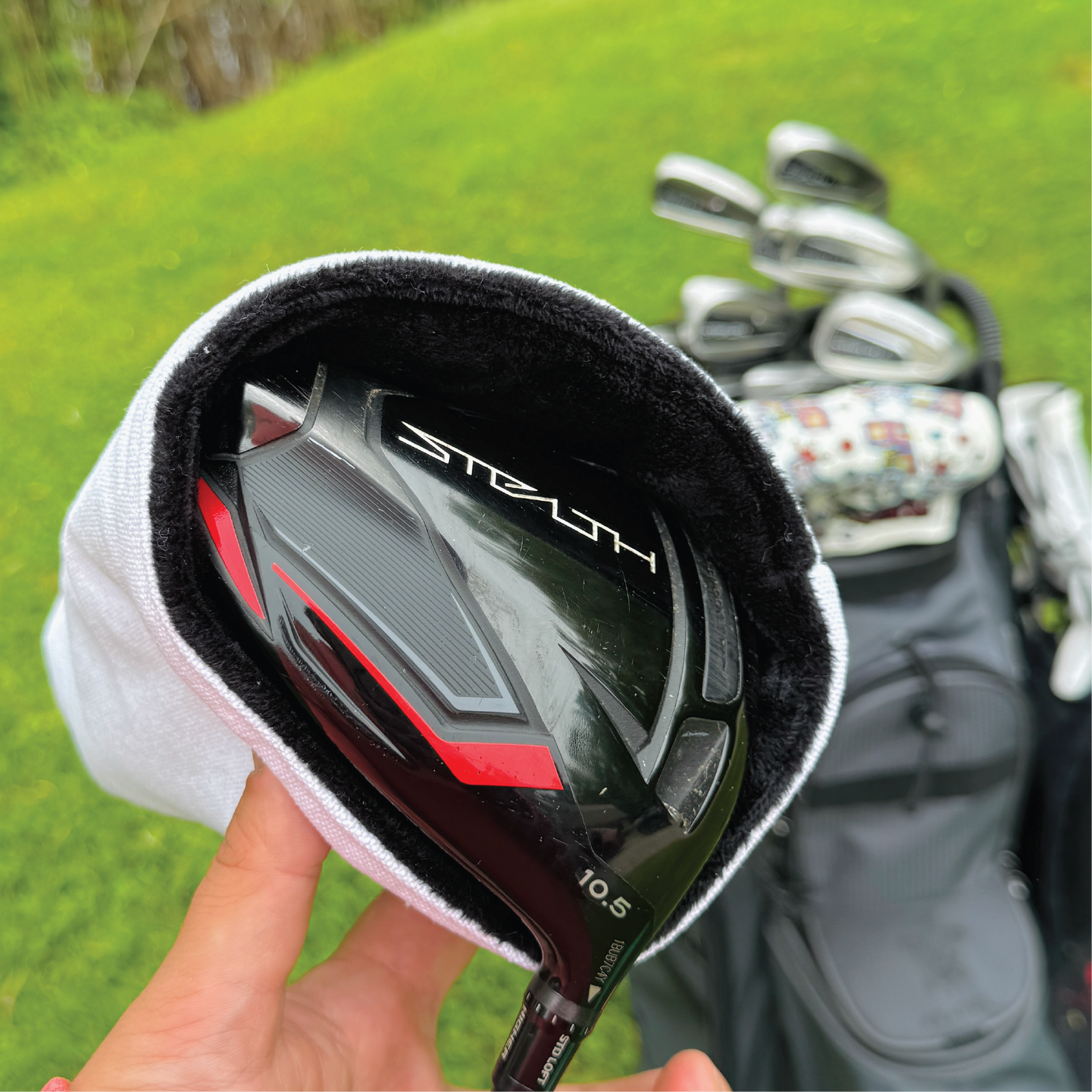 Golf driver with red accents wrapped in white headcover, additional golf clubs and patterned headcover visible on grass background.