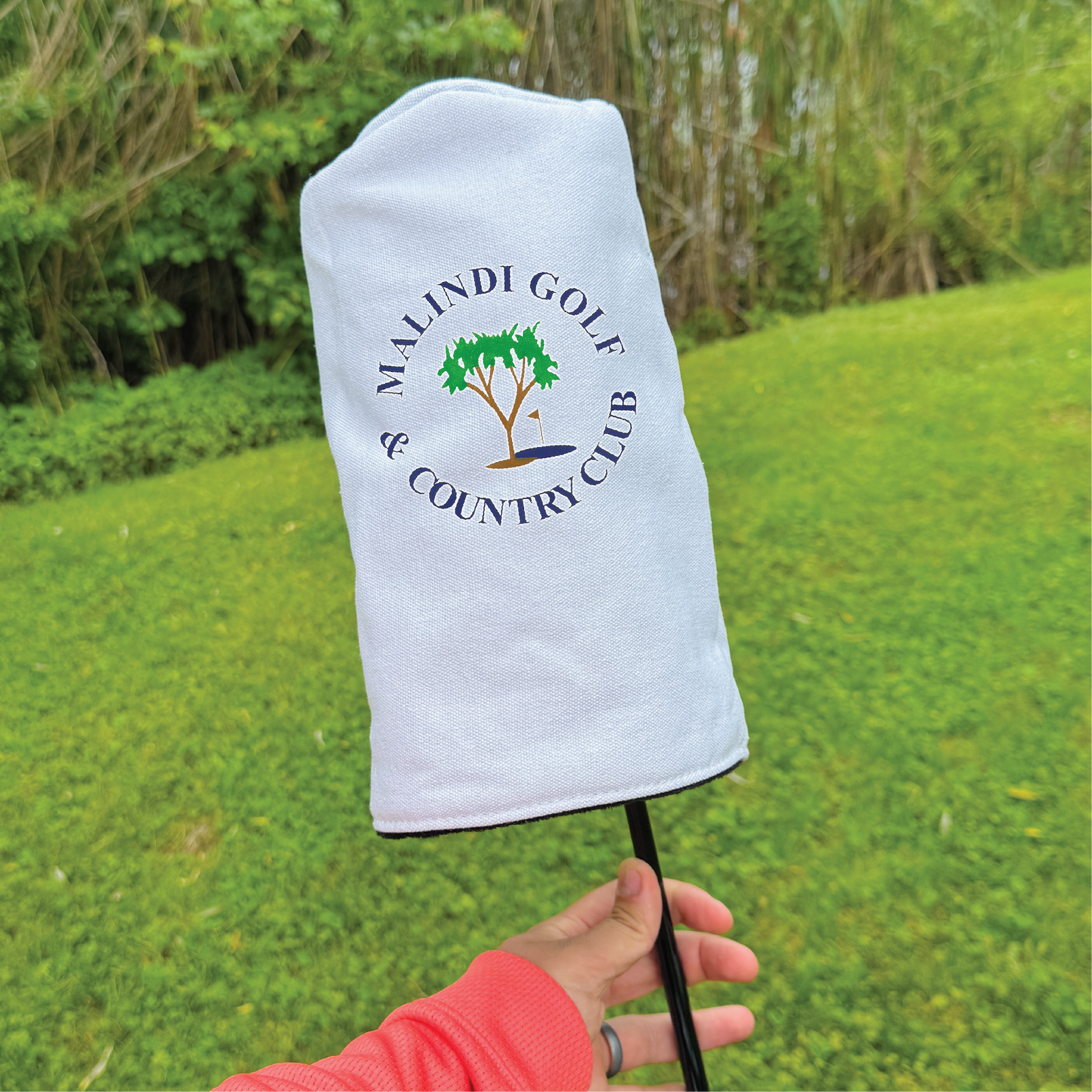 White golf club headcover with embroidered Maldi Golf & Country Club logo featuring a tree and flag design, held on a golf course.