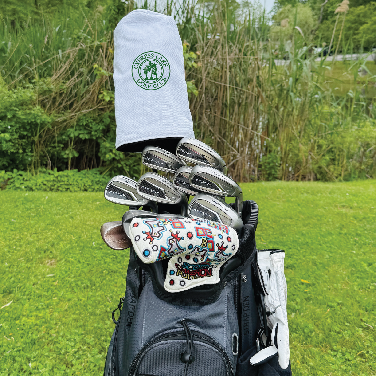 White barrel-style golf headcover embroidered with green Cypress Lake Golf Club logo, displayed on golf bag with irons and colorful putter headcover at golf course.