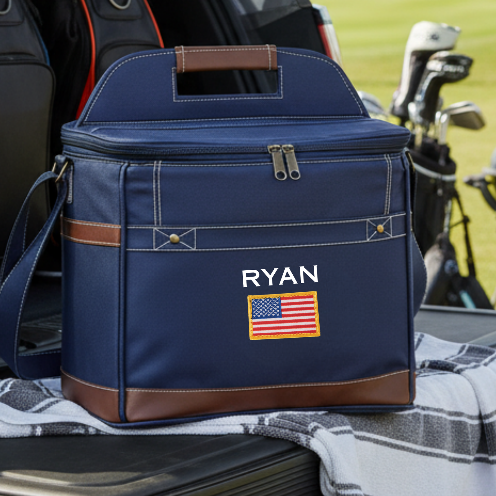 Navy cooler with brown leather trim, brown handle, sample personalization reading "RYAN" with American flag patch, positioned on golf cart with clubs visible in background