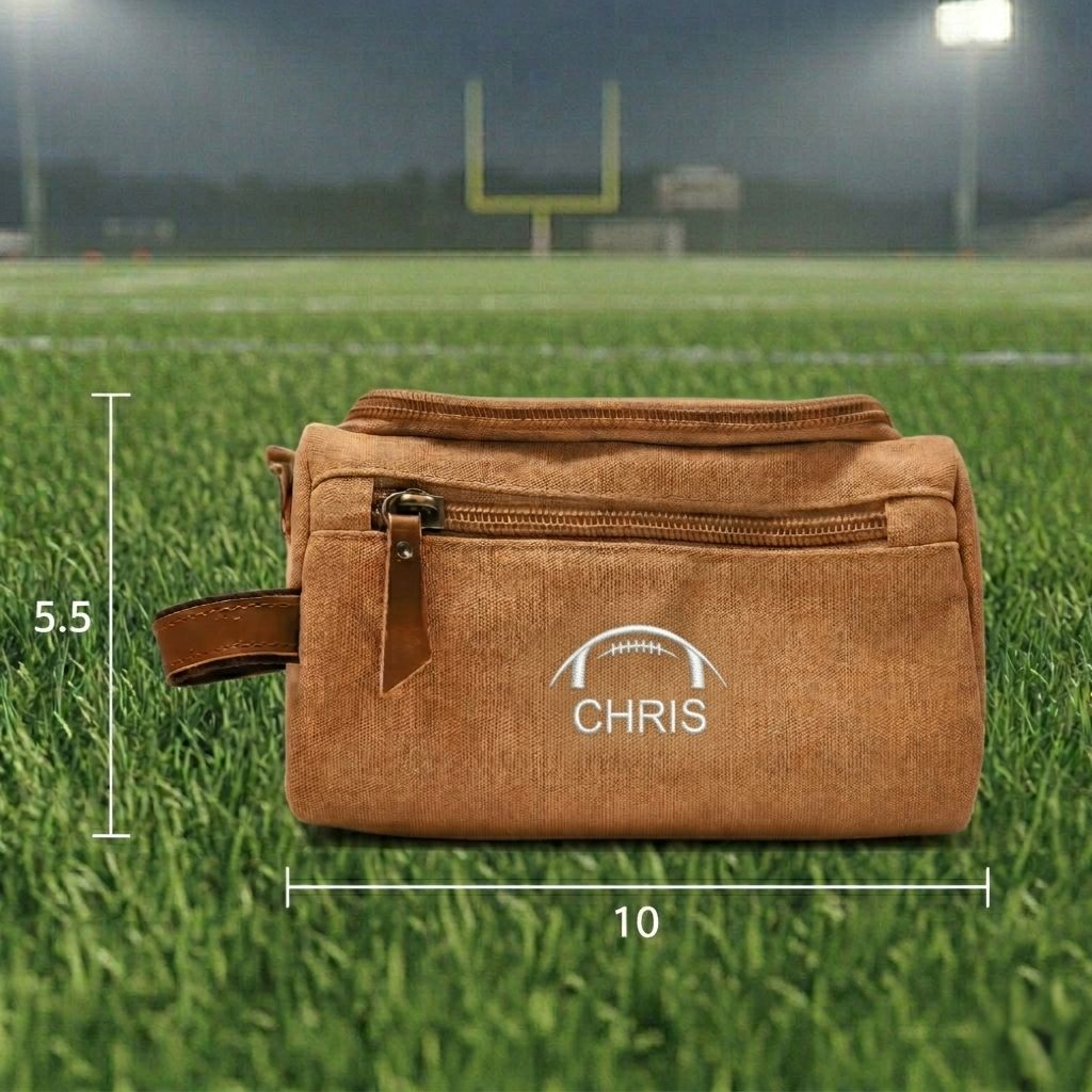 Tan canvas toiletry bag with leather handle, embroidered with sample name "CHRIS" and football design, displayed on football field with goal posts in background, dimensions 10" x 5.5" shown